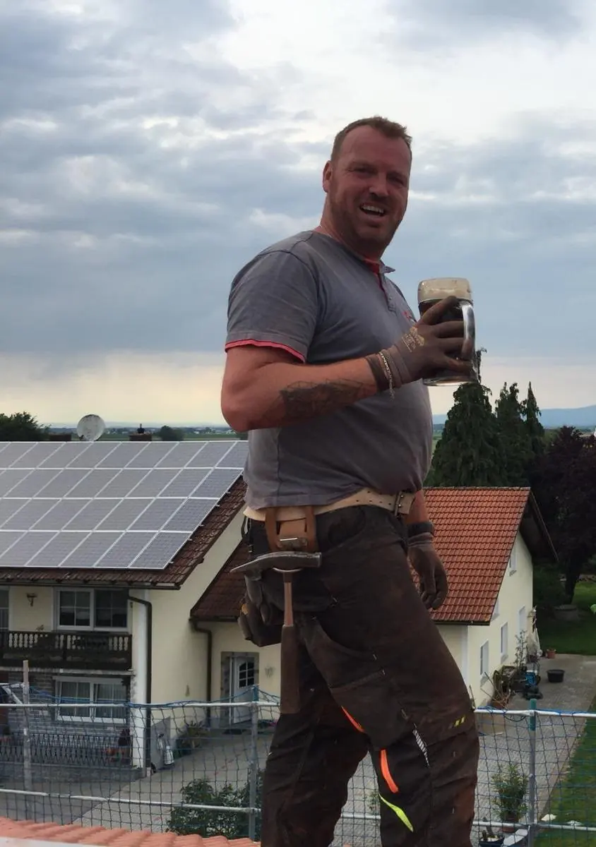Stefan Kärger mit Werkzeuggürtel und einem Glas Weißbier auf einem Dach